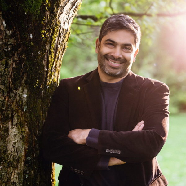 Sirish smiles and poses next to a tree bark. He is standing with his arms folder sporting a dark maroon blazer.