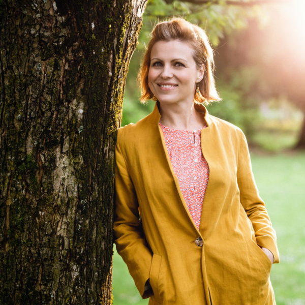 Laura smiles and poses for the camera. She is wearing a mustard blazer with her pink blouse underneath. She leans on the bark of a tree.