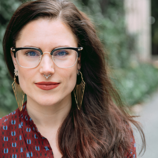 Laura June Albert wears terracotta lipstick while smiling. Laura June is accesorized with large, gold geometric earrings, black half rimmed glasses and a silver septum piercing.