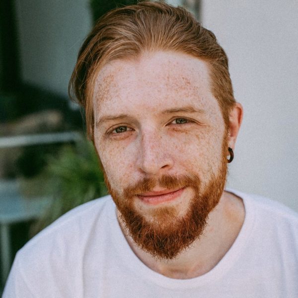 Rob Eccles smiles gently, wearing a white v-neck shirt, face adorned with freckles and a red beard. Rob's red hair is pushed back.