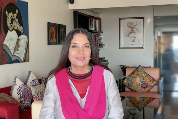 Shabana Azmi smiles shyly at the camera, sitting in a living room. Shabana wears a pale grey kurta, a hot pink dupatta/scarf and assorted jewelry. Shabana has long wavy dark brown hair with some grey streaks. Indian Summer Festival logo in the top left corner.
