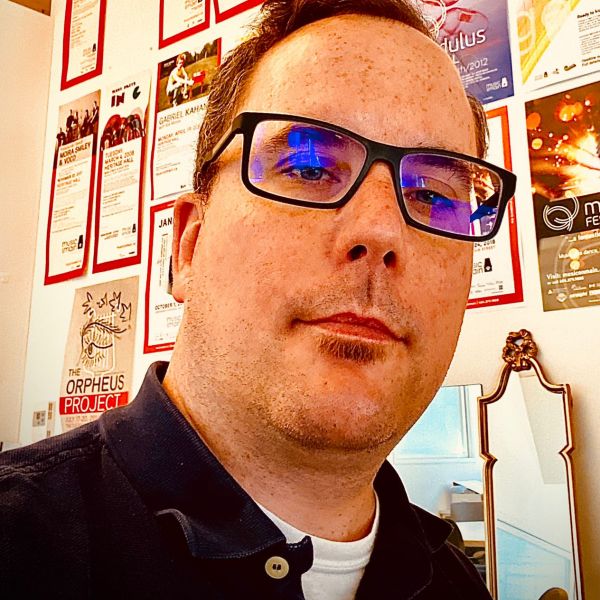 Joseph is facing the camera. He has glasses on. In the background, a small wooden mirror and several poster adorn the wall.