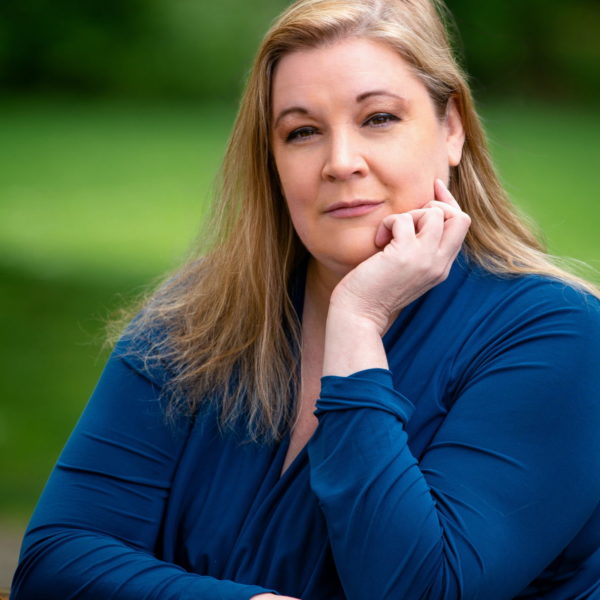 Melissa looks at the camera with her hand resting on her chin. Her straight hair falls on her shoulders.She's sporting a blue blouse. Blurred green background is visible.