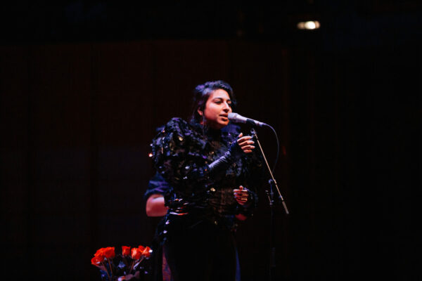 Photo of Arooj Aftab standing on stage singing on a microphone, wearing a black goth style blouse with feathery sleeves.