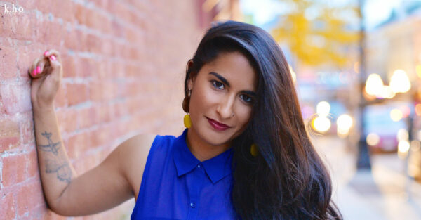 Photo of Anoushka Ratnarajah smiling at the camera, sporting a blue sleeveless collared blouse, standing with one hand resting on the brick wall.