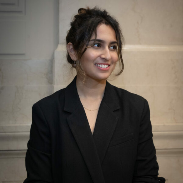 Photo of Sakshi Taneja, with her hair tied up sporting a black blazer and smiling looking away from the camera.