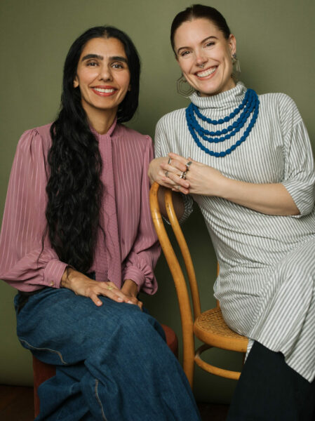 Pawan Deol and Laura June Albert smile at the camera sitting on a wooden chair. Pawan is wearing a flowy pink blouse, her hair falls on her right shoulder and Laura june s wearing a stripped turtleneck dress accessorized with a blue beaded necklace.