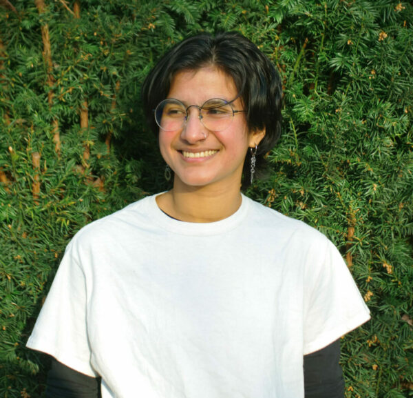 Photo of Tajliya sporting a white tshirt smiling and standing in front of a lush green bush.